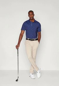 Tamsiai mėlynas polo marškinėlis su baltu raštu, oranžinė apykaklės detalė; derinamas su smėlio spalvos kelnėmis ir baltais golfų batais, laiko golfą lazdą.