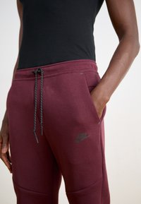 Bordo Nike trenirke s crnim logom, s vezicom u struku, bočnim džepovima i rebrastim materijalom na gležnjevima koji dodaje teksturalni detalj.