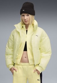 Puma gul pufferjacka med hög krage, dragkedjeslutning och svart logotyp. Matchas med gult joggingbyxor och en svart stickad mössa.