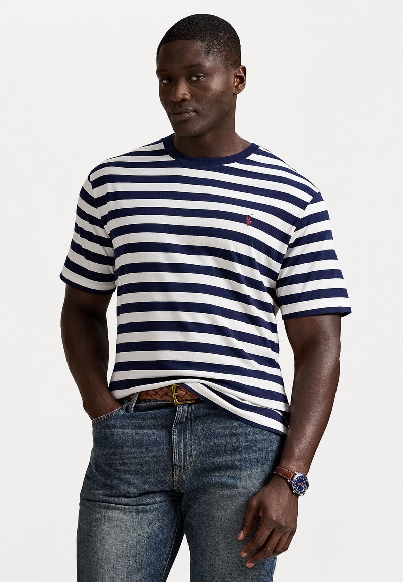 Tumši zils un balts svītrains kokvilnas T-krekls ar apaļu kakla izgriezumu; uz krūšu ir mazs sarkans logotips. Kombinēts ar zilām džinsām un brūnu jostas.