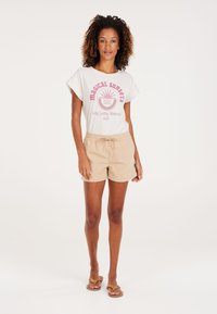 Witte t-shirt met roze grafiek, met de tekst "magische zonsondergangen" en "Langdurige Herinneringen." Beige shorts met elastische tailleband en aantrekkoord.