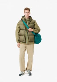Chaleco acolchado verde oliva sobre una chaqueta a juego, combinado con unos pantalones beige y un bolso de hombro en color teal. Zapatillas con detalles en negro y blanco.