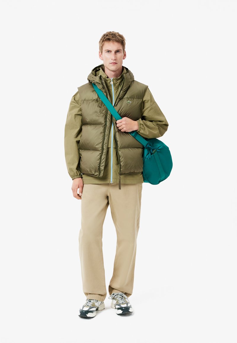 Chaleco acolchado verde oliva sobre una chaqueta a juego, combinado con unos pantalones beige y un bolso de hombro en color teal. Zapatillas con detalles en negro y blanco.