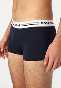 Sötétkék pamut boxer alsónadrág, fehér elasztikus derékpánttal, amely fekete és bézs csíkokkal van díszítve, kiemelve a "BOSS" márkanevet.