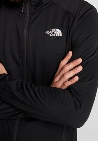 Černá zipová bunda vyrobená z texturovaného materiálu, s malým bílým logem The North Face na levé části hrudi.