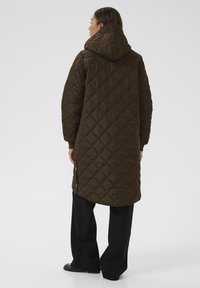 Quilted brun lang frakk med hætte, der har et geometrisk diamantmønster og sprækker i bunden, parret med sorte, vide bukser.