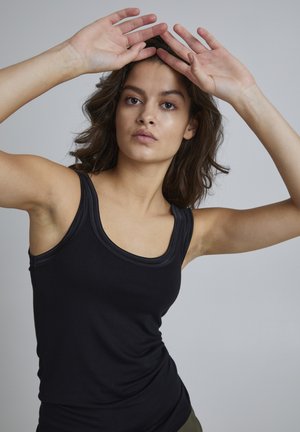 Zwart mouwloos tanktop met een ronde halslijn en textuurafwerking, gemaakt van een soepele, rekbare stof. Model poseert met de handen omhoog.