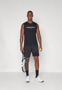 Svart ermeløs skjorte med hvit logo, svarte shorts med lommer, holder en tennisracket og ball, iført hvite treningssko og svarte sokker.