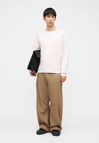Homme portant une chemise à manches longues rose clair, un pantalon beige ample, des chaussures noires, tenant une pochette noire sur un fond blanc uni.