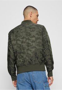 Veste bomber en camouflage sombre avec une texture lisse et brillante. Présente des poignets et un ourlet côtelés, ainsi qu'une silhouette ajustée.