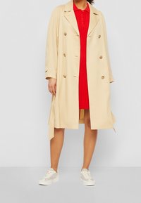 Nainen, jolla on päällään beige trenchcoat punaisen nappimekkonsa päällä, yhdistettynä valkoisiin lenkkareihin, seisoo yksinkertaista vaaleaa taustaa vasten.