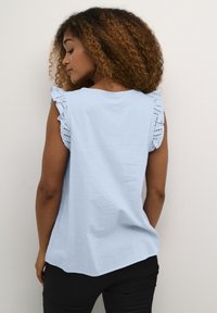 Haut bleu clair à manches courtes avec un ourlet en dentelle découpée et froncée. Tissu doux, coupe décontractée et détail d'ourlet arrondi à l'arrière. Pantalons noirs visibles.