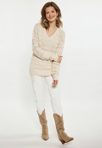 Beige gebreide trui met een V-hals en een textuurpatroon, gecombineerd met witte broek en beige cowboylaarzen met decoratieve stiksels.