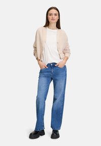 Beige gestrickte Strickjacke mit Knöpfen, weißes T-Shirt und weite blaue Jeans. Schwarze Plateauschuhe. Weiche Textur, lässige Passform.