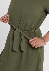 Olivgrön klänning med knytbälte i midjan, som har en mjuk textur och lös passform. Inkluderar flera silverringar på handen.