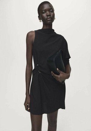 Robe noire au design asymétrique, col montant et détail de nœud sur le côté. Le mannequin tient une pochette foncée. Tissu lisse avec une texture minimale.