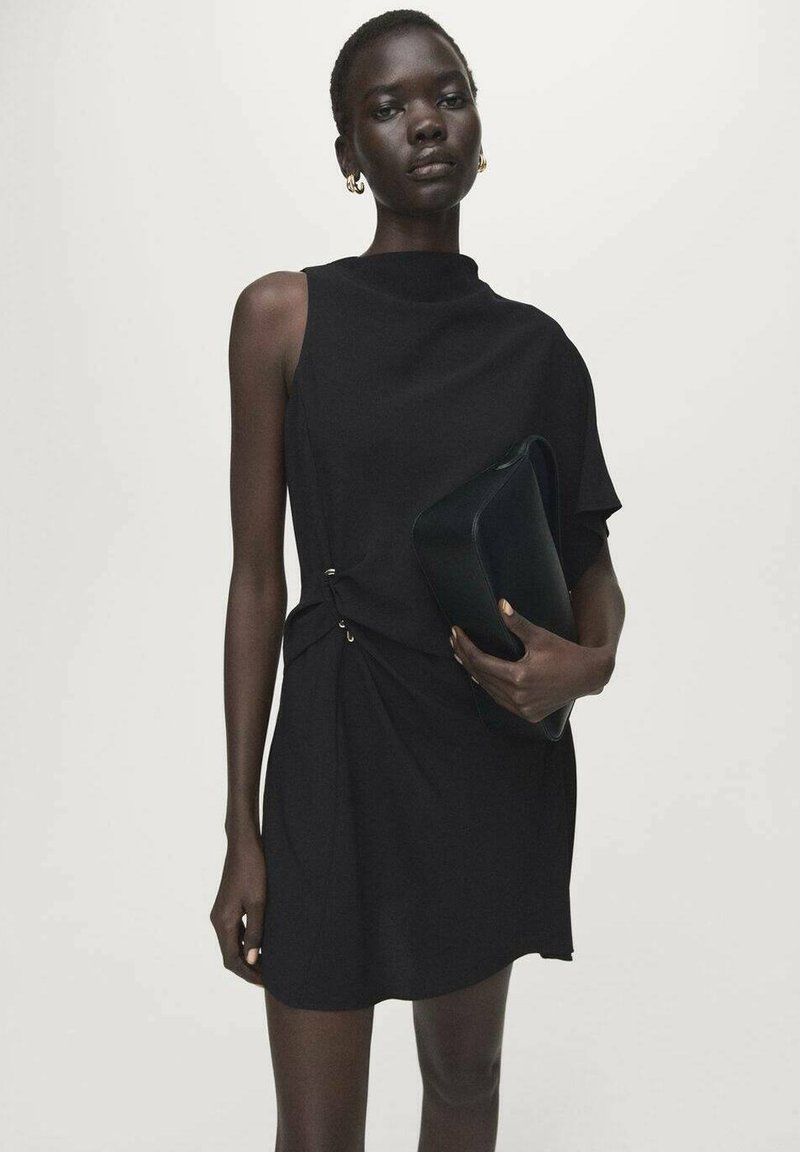 Zwarte jurk met asymmetrisch ontwerp, hoge halslijn en knoopdetail aan de zijkant. Model houdt een donkere clutch vast. Gladde stof met minimale textuur.