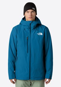 Uomo con una giacca a cappuccio blu The North Face e un berretto nero, in piedi di fronte a uno sfondo bianco uniforme.