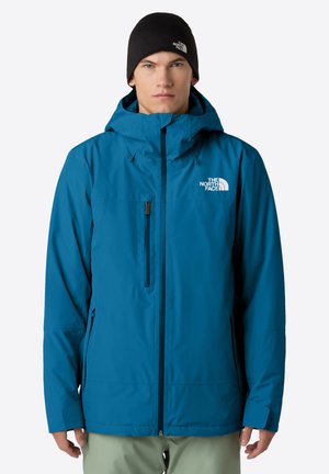 Mężczyzna w niebieskiej kurtce z kapturem The North Face i czapce beanie w kolorze czarnym, stojący na jednolitym białym tle.