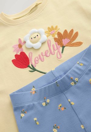 Žluté tričko s vyšívanými barevnými květinami, usměvavou bílou květinou a růžským nápisem "lovely" v kombinaci s modrými kalhotami s malým květinovým potiskem.