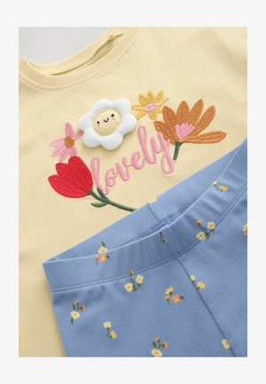 Gul skjorte med broderede farverige blomster, et smilende hvidt blomstermotiv og lyserød tekst "lovely", kombineret med blå bukser med små blomsterprint.