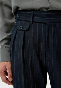 Donkerblauwe gestreepte broek met een hoge taille, voorzien van twee blauwe knopen en een geplooide voorkant, gecombineerd met een grijs shirt.