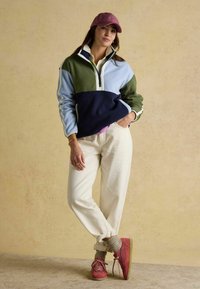 Fleece mikina v modrej, zelenej a navy blokovej farbe, s polovičným zipsom na golieri. Krémové nohavice, ružové tenisky a ružová čiapka dotvárajú outfit.