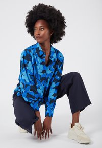 Niebieska koszula w kwiaty z długimi rękawami, noszona z granatowymi spodniami w szerokim kroju i jasnymi sneakersami na białym tle.