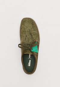 Chaussure en daim vert avec une surface texturée, lacets marron et accent turquoise. Présente un bord cousu et un logo sur la semelle intérieure.
