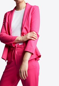 Růžový měkký pletený blazer s šálovým límcem a zapínáním na jeden knoflík, kombinovaný s odpovídajícími kalhotami na šňůrku, které mají hladkou texturu.