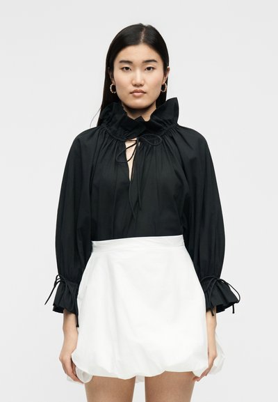 Νεαρή γυναίκα που φοράει μαύρο μπλούζα με μακριά μανίκια, γιακά και μανσέτες με βολάν, σε συνδυασμό με λευκή μίνι φούστα με φουσκωτή ποδόγυρο.