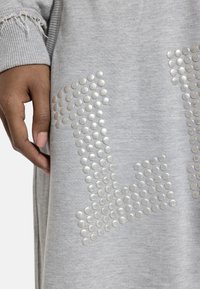 Χέρι που ακουμπάει δίπλα σε γκρι ύφασμα με ασημένιους κυκλικούς καρφίτσες που σχηματίζουν γεωμετρικό μοτίβο, με ορατό φθαρμένο ριμπ μανσέτα.