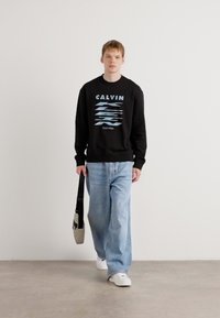 Sweatshirt preto com o texto "CALVIN" em azul, combinado com jeans largos de tom azul claro. Ténis brancos e uma mala de ombro cinza. Fundo minimalista.
