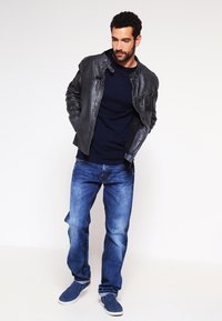 Svart läderjacka över en marinblå tröja, kombinerad med ljusblå denimjeans och marinblå slip-on skor. Modellen står med en avslappnad hållning.