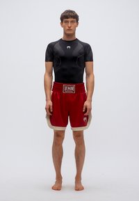 Μαύρο συμπιεστικό πουκάμισο με κοντά μανίκια, σε συνδυασμό με κόκκινα σορτς που έχουν λευκή ζώνη μέσης και λογότυπο. Το μοντέλο στέκεται ξυπόλυτο σε ουδέτερο φόντο.