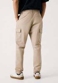 Beige cargobroek met zijzakken en een slanke pasvorm, gemaakt van duurzaam materiaal. Gepaard met witte sneakers met een bruine zool.