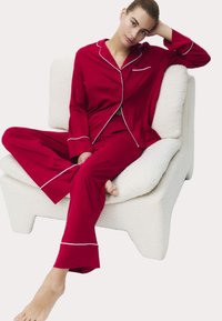 Rød pyjamas sæt med hvid piping; har lange ærmer, knaplukning foran og løstsiddende bukser. Model siddende på en tekstureret creme farvet stol.