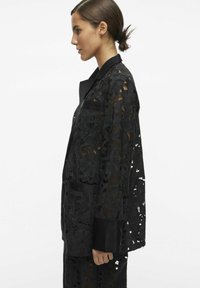 Veste en dentelle noire brodée avec des motifs floraux, un col et des manches longues, agrémentée d'accents en tissu noir uni.