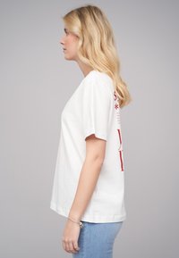 T-shirt en coton blanc à manches courtes, avec du texte et des graphiques rouges au dos. Le modèle porte un jean bleu clair.