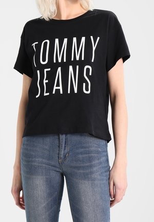 Osoba ubrana w czarny krótki rękaw T-shirt z napisem "TOMMY JEANS" na białych literach oraz niebieskie dżinsy.