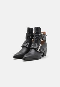 Las botas de cuero negro hasta el tobillo presentan una punta afilada, un tacón cuadrado y correas decorativas con herrajes de plata y ojales. Textura suave y diseño estructurado.