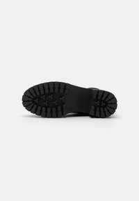 Suela de bota de goma negra con surcos profundos para tracción, con un logotipo de la marca grabado en el lateral y un tacón liso.
