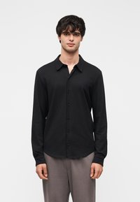 Μαύρο πουκάμισο μακρυμάνικο με γιακά με κουμπιά, κατασκευασμένο από λείο ύφασμα. Διαθέτει ευθεία άκρη και διακριτικό μπροστινό κούμπωμα με κουμπιά.