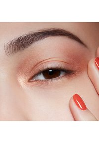 Gros plan sur un œil avec un fard à paupières bronze chaud, un sourcil bien défini et une manucure rouge brillante. La peau a une texture lisse ; l'œil est marron avec des cils visibles.