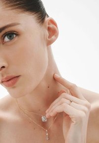 Collares de oro con un colgante rectangular y un charm en forma de lágrima, llevados con un delicado anillo en un dedo, resaltando la textura de la piel.
