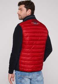 Gilet rosso imbottito con cuciture orizzontali, maniche nere e colletto blu navy. Presenta logo e testo sulla parte superiore della schiena. Tessuto testurizzato.