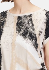 Luźna bluzka w odcieniach kremu, czerni i beżu z abstrakcyjnym wzorem plam farby. Miękka tkanina, okrągły dekolt i krótkie rękawy.