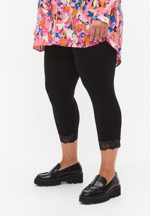 Zwarte capri legging met kanten rand aan de zoom, gecombineerd met glanzende zwarte loafers. De top is een kleurrijke, gestreepte blouse.