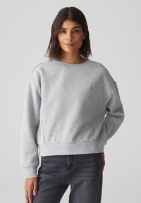 Grauer Pullover mit Rundhalsausschnitt, abgesenkten Schultern und gerippten Bündchen. Verfügt über seitliche Nahttaschen und ein kleines Logo auf der Brust.