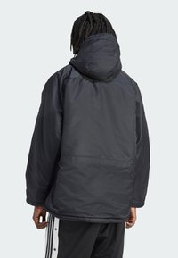 Person med mørkt flet hår iført en sort hættetrøje og sorte bukser med hvide side-striber, vendende væk mod en ensfarvet baggrund.